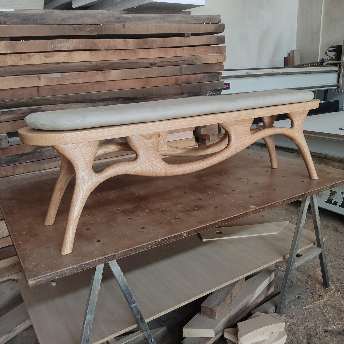 May include: A light-colored wooden bench with a curved design and a padded seat. The bench has a unique, open-frame structure with curved legs and a light-colored cushion. The bench is likely made of oak or a similar hardwood.