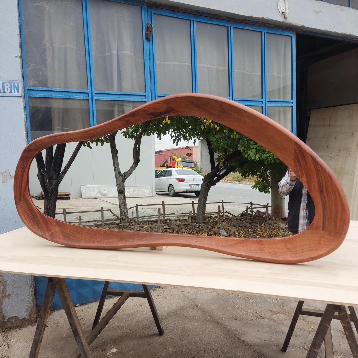 May include: A large, organically shaped wooden mirror with a rich, reddish-brown finish. The mirror reflects a view of trees, a white car, and a building. The mirror is displayed on a light-colored wooden surface.