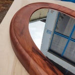 May include: Oval-shaped wooden mirror with a rich, dark brown frame. The frame has a smooth, curved design. The mirror reflects a view of a building with blue window frames and a cloudy sky.
