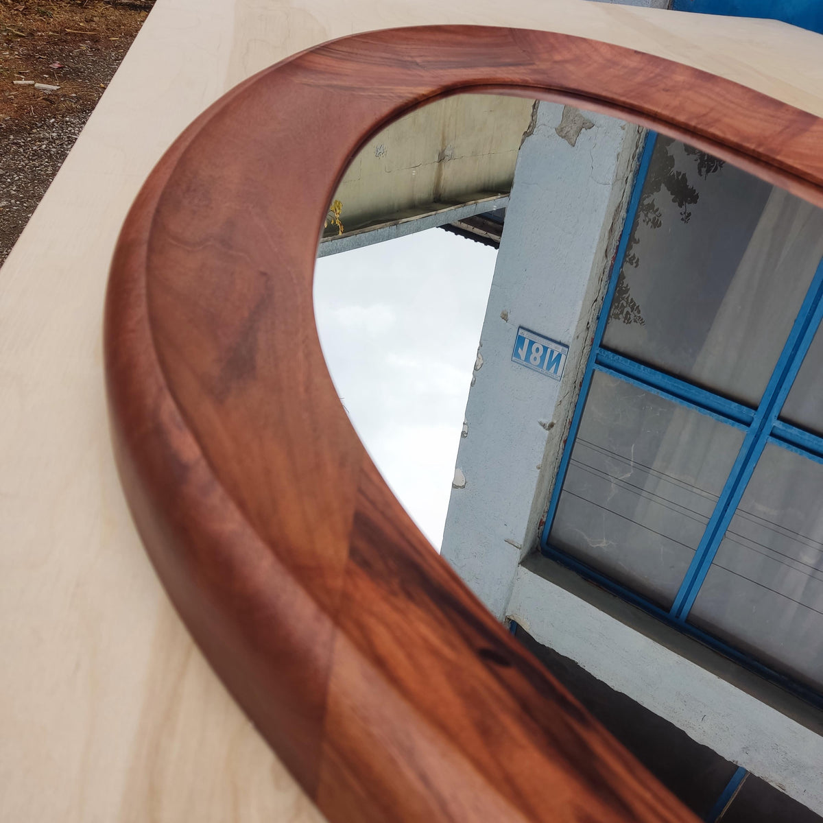May include: Oval-shaped wooden mirror with a rich, dark brown frame. The frame has a smooth, curved design. The mirror reflects a view of a building with blue window frames and a cloudy sky.