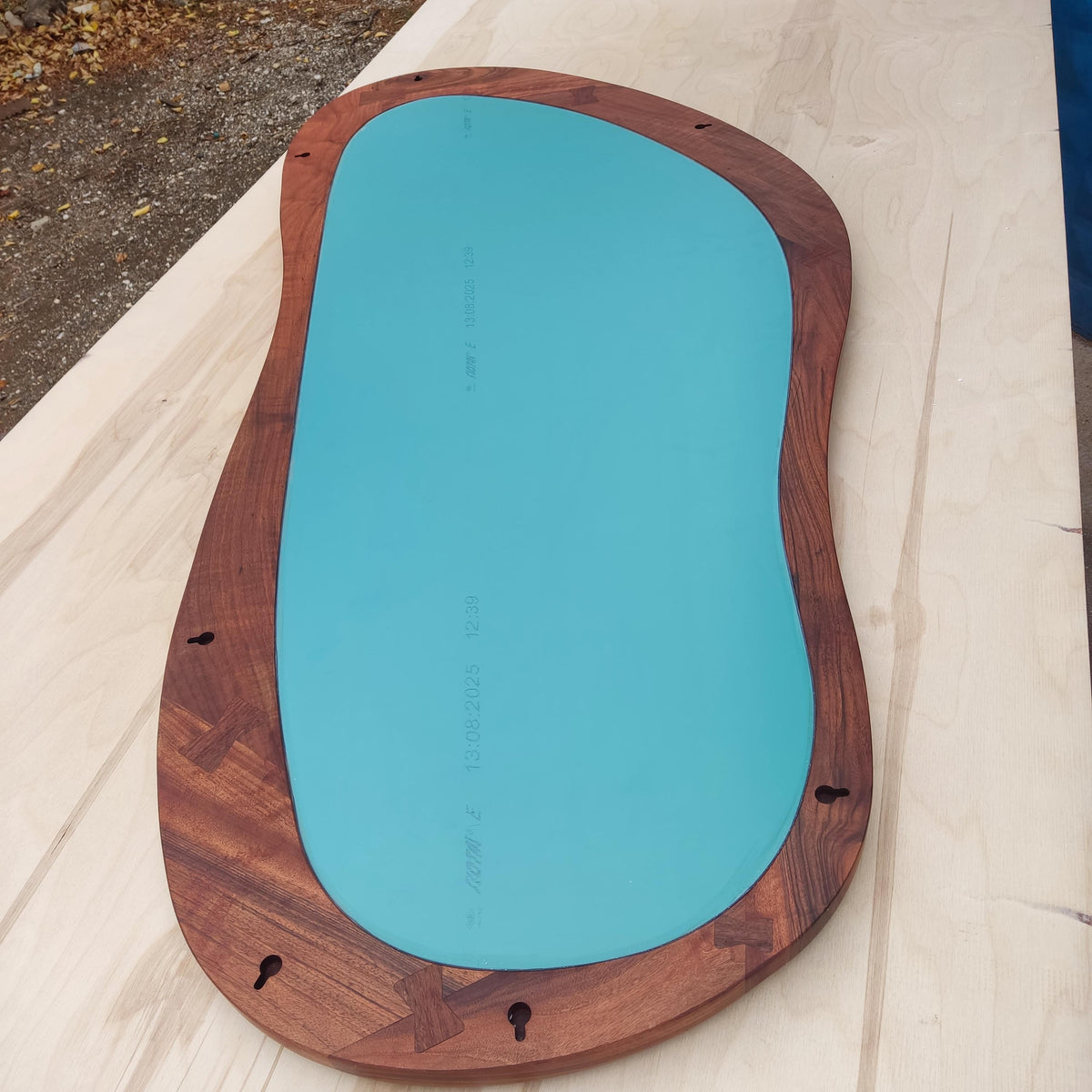 May include: A modern, organic-shaped mirror with a teal-colored glass surface. The mirror is framed with dark brown wood, featuring decorative joinery at the corners. Includes keyhole hangers for wall mounting.