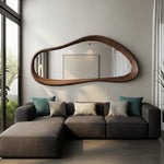 May include: A modern, organic-shaped mirror with a dark wood frame hangs above a gray sectional sofa. The mirror's unique design adds a touch of elegance to the living room. Decorative pillows in teal, gray, and beige complement the sofa's neutral tones.