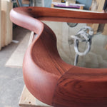 May include: Close-up of a curved, wooden table edge. The wood is a rich, reddish-brown color with visible grain. The design features a smooth, flowing curve, suggesting a modern or mid-century style. The table appears to have a glass top.