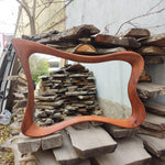May include: A large, wavy-edged mirror with a reddish-brown wooden frame. The frame has a unique, flowing design. The mirror is displayed outdoors, resting on a pile of wooden planks and logs, suggesting a rustic or handcrafted aesthetic.