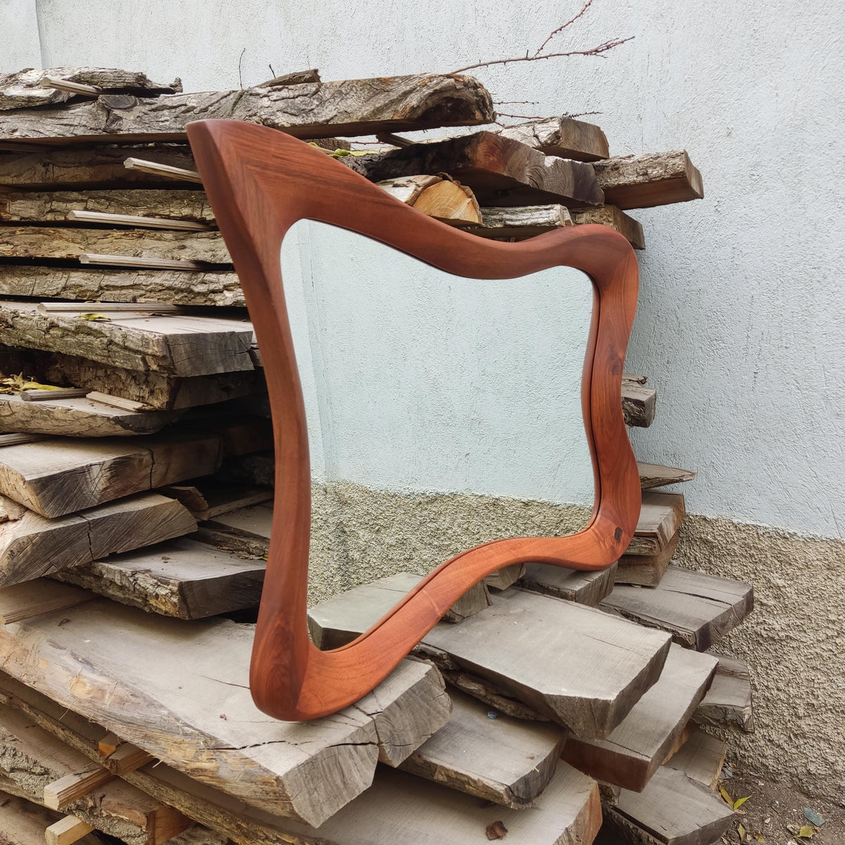 May include: A wooden mirror with a unique, wavy frame. The frame is a warm reddish-brown color and surrounds a clear mirror. The mirror is displayed against a background of stacked wooden planks.