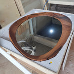 May include: Oval-shaped wooden mirror with a dark brown, natural wood grain frame. The mirror is set in a white protective frame, reflecting a room with a bright light source. The mirror's unique shape and wood grain add a natural, rustic aesthetic.