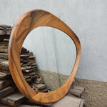 May include: Oval-shaped wooden mirror with a unique, flowing frame. The frame is made of light brown wood with dark brown grain patterns. The mirror reflects a gray wall and is set against a background of stacked wooden planks.