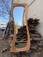 May include: A full-length floor mirror featuring a tall, organic, wavy wooden frame with a natural light brown wood grain finish. The mirror has a smooth, continuous curved shape that creates an elongated, irregular opening for the reflective glass surface.