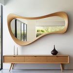 May include: A large, organically shaped wooden mirror hangs above a wooden console table. The mirror's frame has a flowing, wavy design. The console features three drawers and tapered legs. Two white vases and a dark brown vase sit on the console.