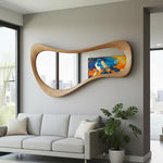 May include: A large, modern, wooden mirror with a unique, flowing shape. The mirror is mounted on a gray wall above a gray sofa. An abstract painting with blue, yellow, and orange hues is visible in the mirror's reflection.