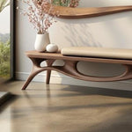 May include: A modern wooden bench with a curved design and a light-colored cushion. The bench has a unique shape with cut-out details and is paired with a white vase, a small decorative sphere, and a mirror.
