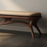 May include: A modern wooden bench with a light brown, cushioned seat. The bench has a dark brown, organically shaped base with curved legs. The bench is set against a neutral wall and floor.