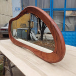 May include: A large, organically shaped mirror with a rich, reddish-brown wooden frame. The mirror is positioned horizontally on a light-colored wooden surface, reflecting the outdoor environment. The frame has a smooth, curved design.