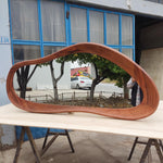 May include: A large, organically shaped wooden mirror with a rich, reddish-brown finish. The mirror reflects a view of trees, a white car, and a building. The mirror is displayed on a light-colored wooden surface.