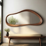 May include: A modern, organic-shaped wooden mirror with a built-in shelf hangs above a wooden bench with a cream-colored cushion. A black vase with greenery sits on the bench. The mirror reflects a tree.