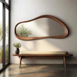 May include: A modern, organic-shaped wooden mirror hangs above a wooden bench with a cream-colored cushion. A black vase with greenery sits on the bench. The mirror reflects a tree and the room's natural light.
