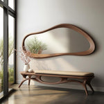May include: A modern entryway scene featuring a large, organically shaped wooden mirror above a matching bench. The bench has a light-colored cushion. A white vase with pink flowers and a small white decorative object sit on the bench. The room has a large window.