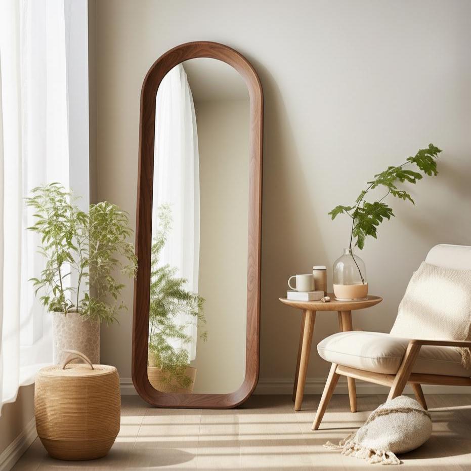 May include: A tall, arched-top, full-length mirror with a dark brown wooden frame. The mirror is placed against a neutral-colored wall, reflecting a bright window and a room with a chair and side table. The mirror is a decorative home decor item.