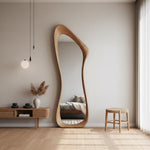 May include: A large, free-form wooden mirror leans against a neutral-toned wall. The mirror's frame has a unique, organic shape. A small, round wooden stool and a low, wooden cabinet with decorative items are also in the room. The floor is made of light-colored wood.