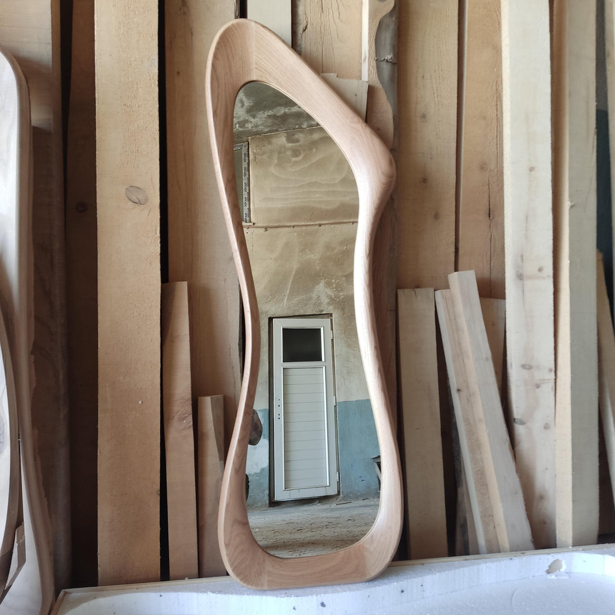 May include: A full-length, free-form wooden mirror with a light brown finish. The mirror's frame has a unique, flowing shape. The background shows wooden planks and a glimpse of a room with a white door.