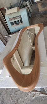 May include: A wooden framed mirror with a unique, flowing shape. The frame is made of light brown wood, and the mirror reflects the surrounding workshop environment. The mirror is resting on a white surface.