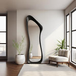 May include: A tall, black, full-length mirror with an organic, flowing shape. The mirror leans against a white wall in a modern living space. A white rug, a beige armchair, and potted plants complete the room's decor.