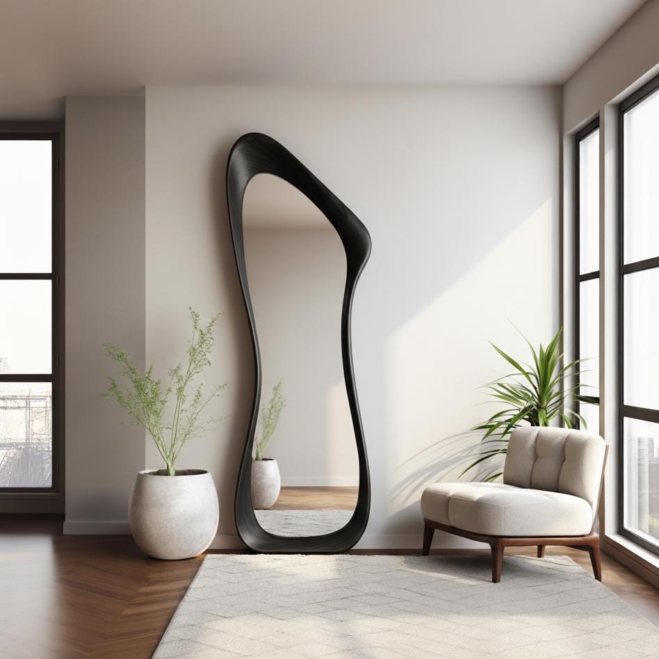 May include: A tall, black, full-length mirror with an organic, flowing shape. The mirror leans against a white wall in a modern living space. A white rug, a beige armchair, and potted plants complete the room's decor.