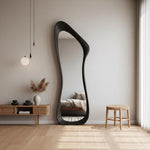 May include: A large, black, organically shaped floor mirror. The mirror has a unique, flowing silhouette and is set against a neutral-toned wall. The room also features a wooden side table, a small stool, and a decorative vase with dried plants.