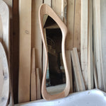 May include: A tall, uniquely shaped wooden mirror with a light brown frame. The mirror has an organic, flowing design, with a reflective surface. The background features various wooden planks and beams, suggesting a workshop or construction setting.