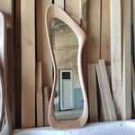 May include: A full-length, free-form wooden mirror with a light brown finish. The mirror's frame has a unique, flowing shape. The background shows wooden planks and a glimpse of a room with a white door.