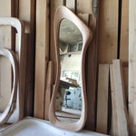 May include: A light-colored, wooden, free-form mirror with a curved, organic shape. The mirror reflects a room with a window and a table. The frame is made of light-colored wood, and the overall design is modern and artistic.