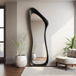 May include: A tall, black, full-length mirror with an organic, flowing shape. The mirror leans against a white wall, reflecting the room. The frame is a solid, dark color. The room features a white rug, a beige armchair, and potted plants.