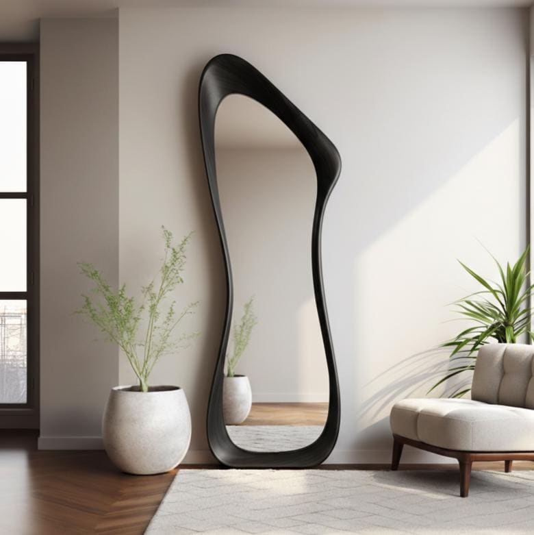 May include: A tall, black, full-length mirror with an organic, flowing shape. The mirror leans against a white wall, reflecting the room. The frame is a solid, dark color. The room features a white rug, a beige armchair, and potted plants.