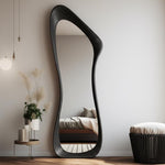 May include: A full-length, black, organically shaped mirror leans against a white wall. The mirror's frame has a unique, flowing design. The reflection shows a bedroom with a bed and a small side table with plants. A black stool sits nearby.