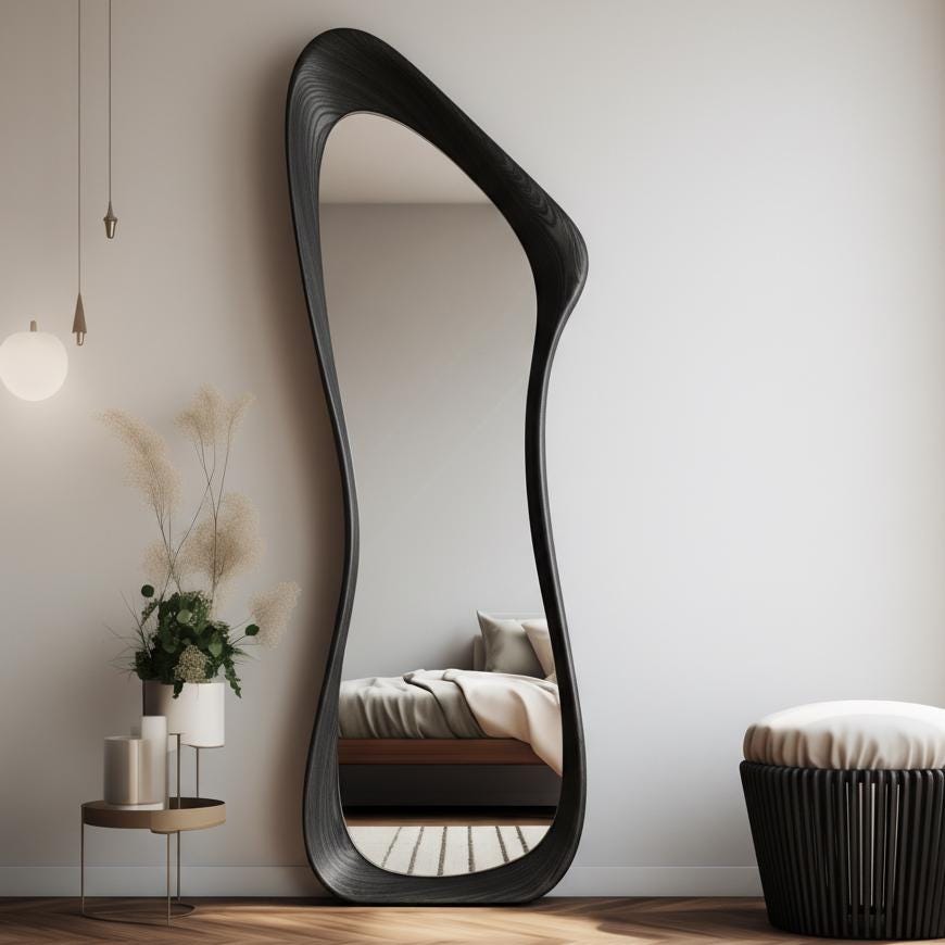 May include: A full-length, black, organically shaped mirror leans against a white wall. The mirror's frame has a unique, flowing design. The reflection shows a bedroom with a bed and a small side table with plants. A black stool sits nearby.