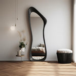 May include: A large, black, full-length mirror with an organic, flowing shape. The mirror leans against a white wall, reflecting a bedroom scene. A small side table with plants and candles sits nearby, along with a black stool with a white cushion.