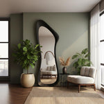 May include: A large, black-framed, full-length mirror with an organic shape stands against a sage green wall. The mirror reflects a living room with a beige sofa, a small table, and a white armchair. A potted plant sits on a woven rug.