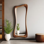 May include: A tall, wooden-framed mirror with a unique, flowing shape. The mirror is made of a warm-toned wood and leans against a light gray wall. The reflection shows a small plant in a white pot. A matching wooden stool with a tan cushion is also visible.
