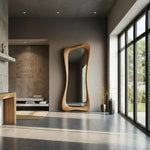 May include: A tall, wooden-framed mirror with an organic, flowing shape. The mirror is mounted on a neutral-toned wall in a modern interior space. The room features large windows, a gray tiled floor, and minimalist decor.