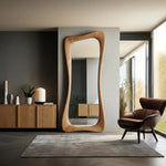 May include: A large, free-standing, wooden mirror with an organic, flowing shape. The mirror is made of light-colored wood and stands tall against a neutral-colored wall. The room also features a wooden cabinet, a rug, and a leather armchair.