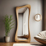 May include: A full-length, free-standing mirror with a unique, organic wooden frame. The mirror's shape is asymmetrical, with gently curving edges. The wood has a natural, light brown finish. The mirror reflects a room with a bed and a window.