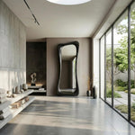 May include: A modern, elongated, black-framed mirror with an organic, wavy shape dominates a minimalist interior. The room features a neutral color palette, with concrete walls and flooring, and a large window overlooking a garden. Decorative items are placed on floating shelves.