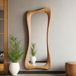 May include: A tall, wooden-framed mirror with a unique, flowing shape. The light-colored wood grain is visible. The mirror is mounted on a gray wall, reflecting a small plant in a white pot. A matching wooden stool is visible.
