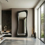 May include: A tall, dark-framed mirror with an organic, flowing shape. The mirror is positioned against a neutral-toned wall in a modern interior. The room features a minimalist design with natural light and decorative vases.