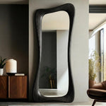 May include: A tall, black, full-length mirror with a unique, organic shape. The frame is made of dark wood and has a flowing, curved design. The mirror is positioned against a neutral-colored wall, reflecting the interior space. The overall aesthetic is modern and minimalist.