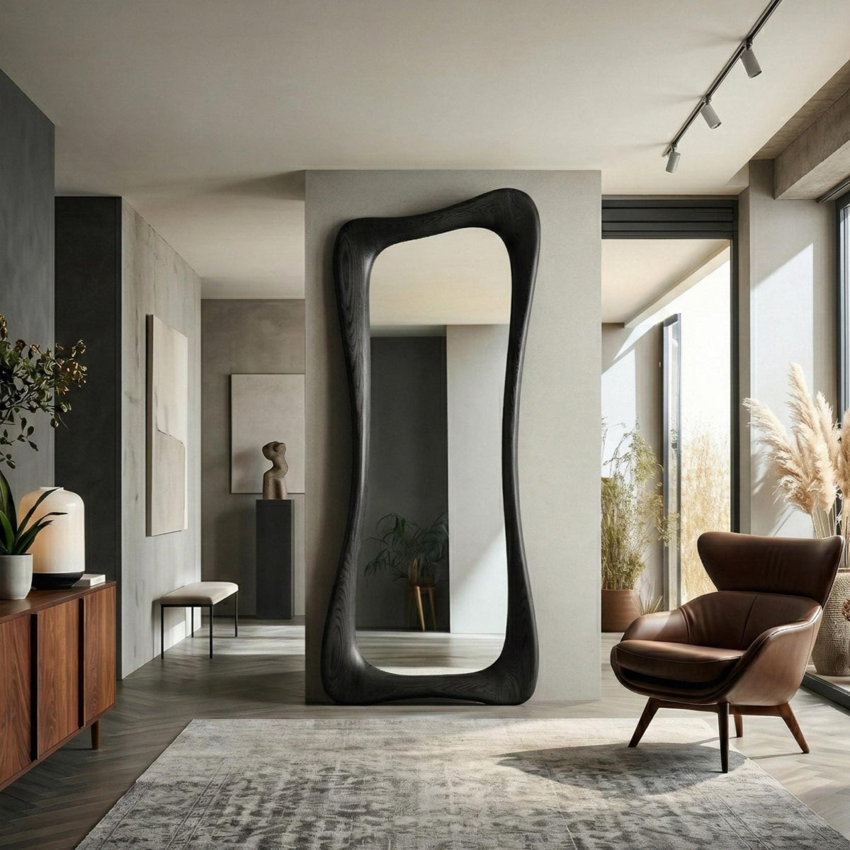 May include: A large, dark-stained wooden mirror with an organic, flowing shape. The mirror is tall and narrow, with a curved frame. The mirror is placed in a modern living space with a brown leather armchair and a gray rug.