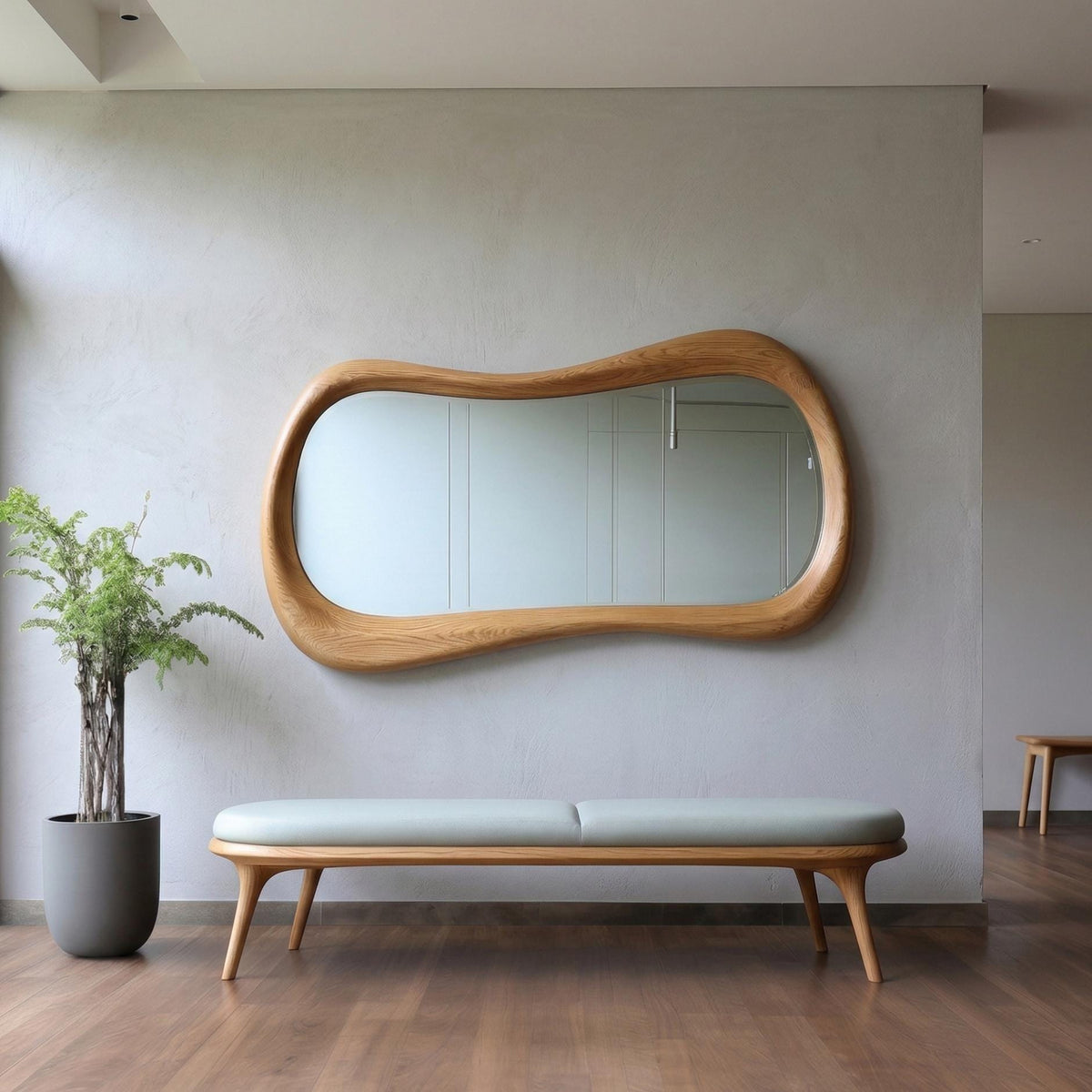 May include: A wall-mounted mirror with an irregular, organic-shaped light wood frame. Below the mirror sits a long bench featuring a light gray upholstered seat supported by a light wood base with four tapered legs. A potted green plant stands nearby.