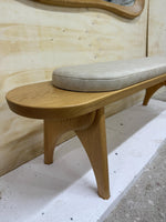 May include: A light-colored wooden bench with a curved design. The bench has a light gray, cushioned seat. The legs are curved and angled, providing support. The bench is made of wood and has a natural finish.