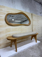 May include: A wooden bench with a light gray cushioned seat and a matching organic-shaped mirror. The mirror has a wooden frame and is mounted above the bench. The bench and mirror are set against a light-colored wooden wall.