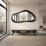 May include: A modern entryway featuring a large, organically shaped black mirror above a matching bench with a beige cushion. The room has a neutral color palette with a large window and a minimalist design.